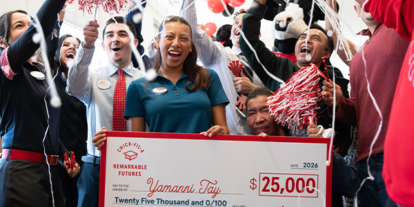 Person smiling and holding a large check, with other people cheering behind her