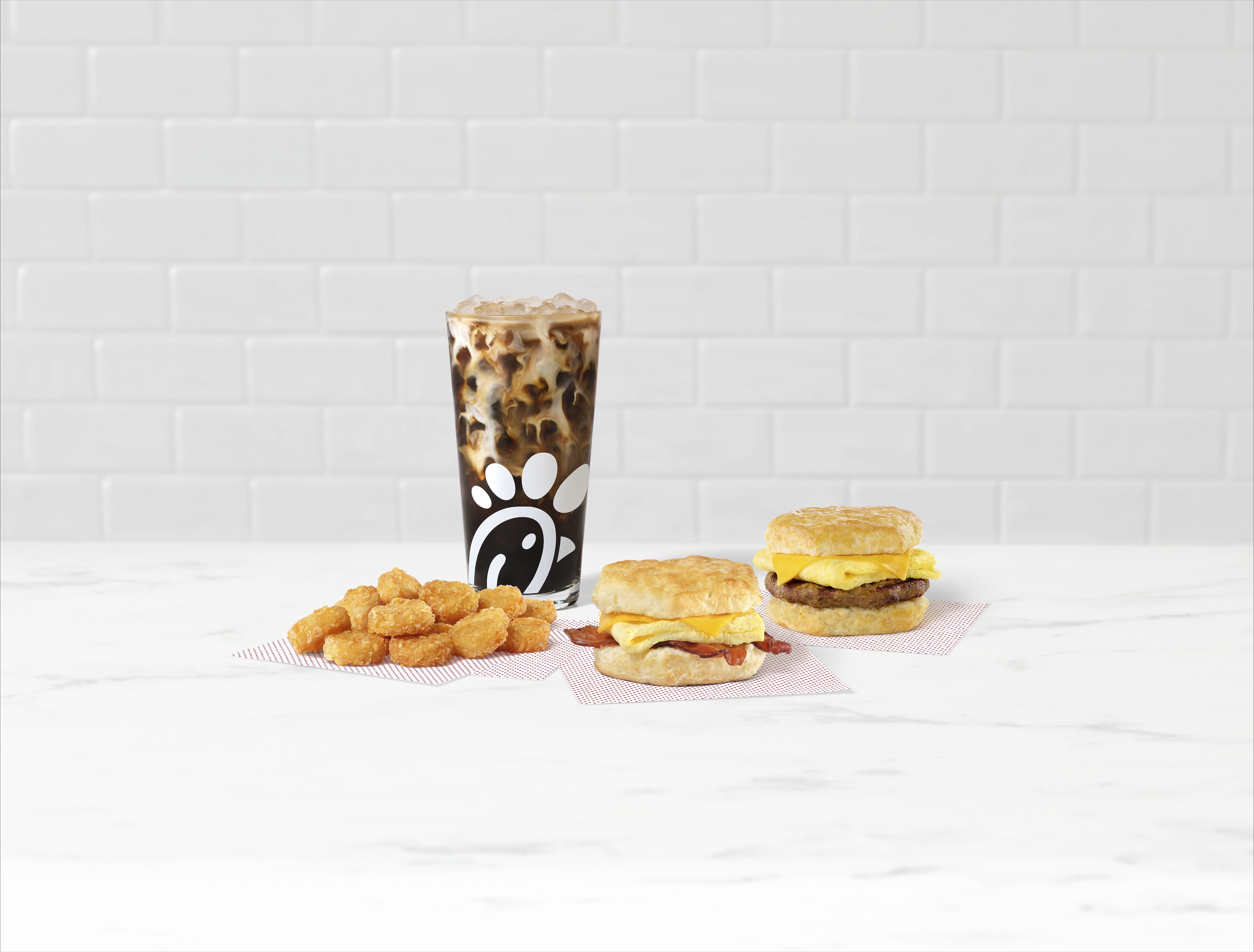 Bacon, Egg & Cheese Biscuit and Sausage, Egg & Cheese Biscuit on parchment paper with Hash Browns on parchment paper and an Iced Coffee on a white counter