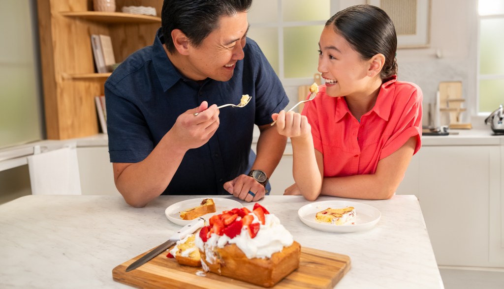 An adult and child smiling at each other and standing together behind a counter with a Strawberry Shake Cake and plates sitting on it 