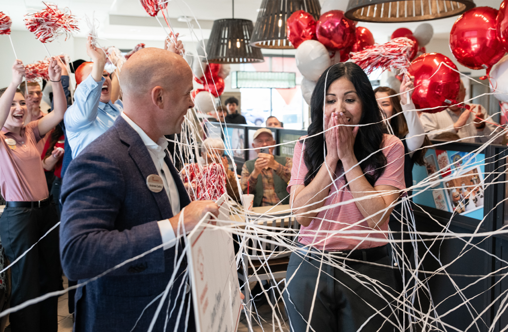 A person handing a check to another person with balloons, silly string and other people cheering around them