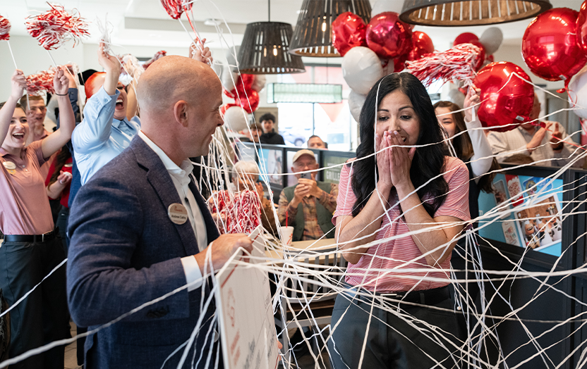 A person handing a check to another person with balloons, silly string and other people cheering around them