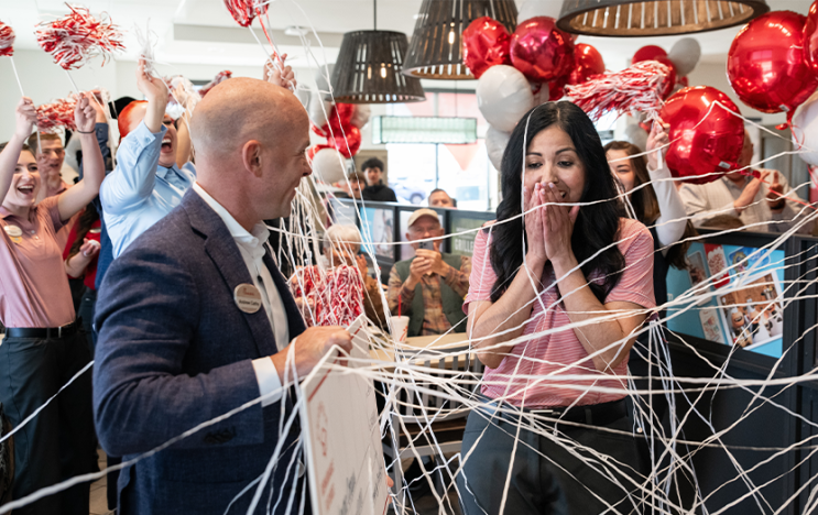 A person handing a check to another person with balloons, silly string and other people cheering around them