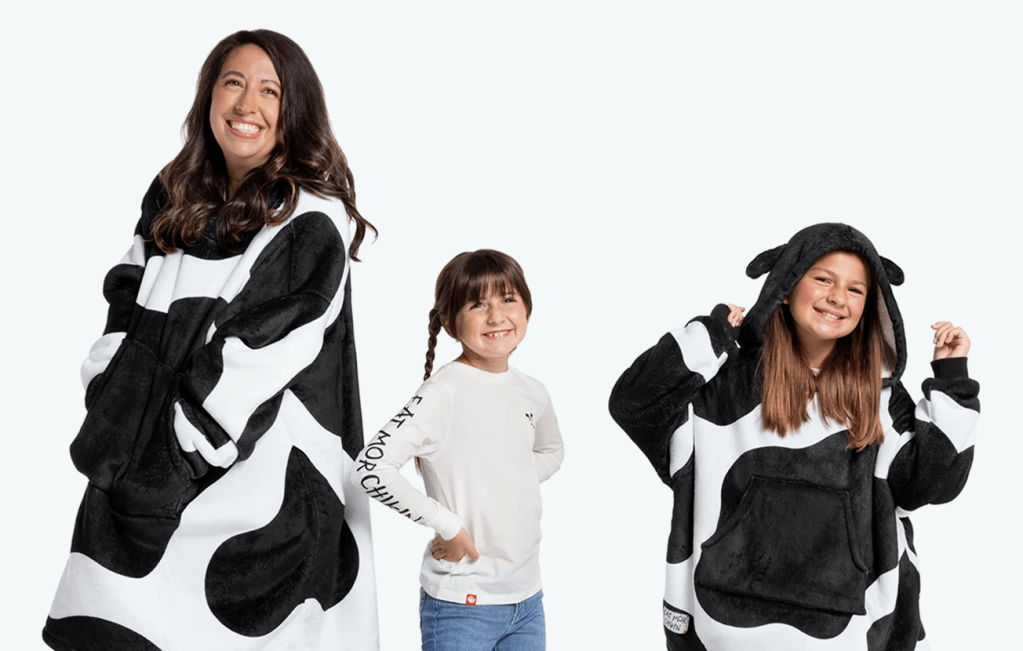 An adult and child wearing a Cow Print Fleece Lined Wearable Blanket with another child in between them wearing a Eat Mor Chikin® Kids Graphics Long Sleeve Shirt