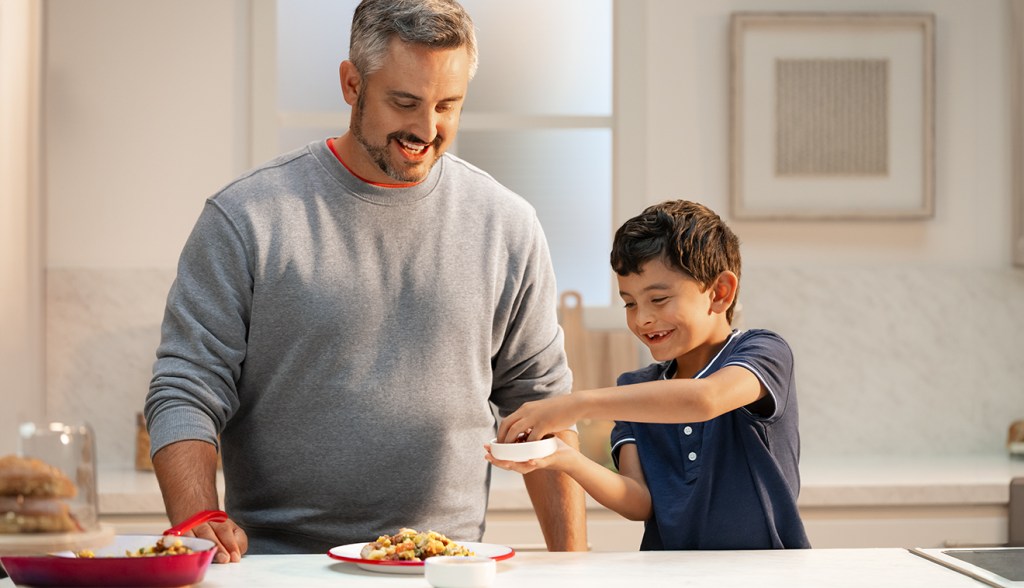 An adult and a child standing together behind a counter with a plate a food, while the child holds a small dish