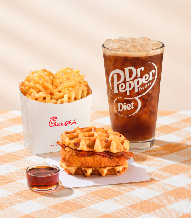 Chicken & Waffles Sandwich on napkins with a side of Classic Syrup next to Waffles Fries and a glass of Diet Dr Pepper on a brown checkered tablecloth