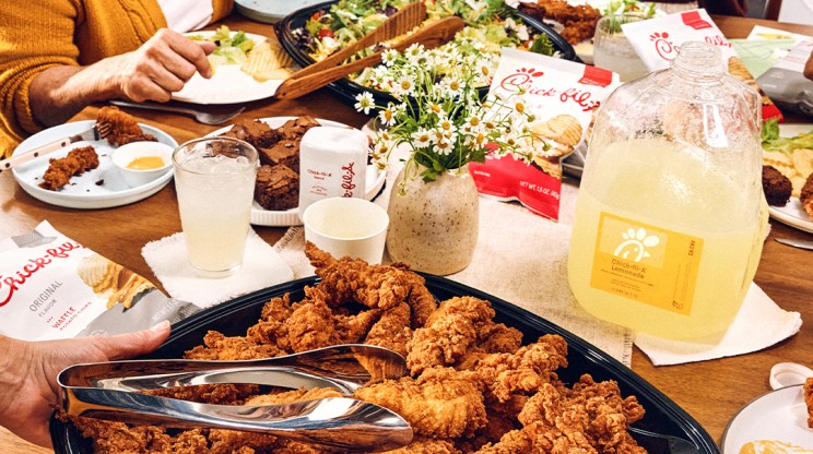 People gathered around a table with a Chick-n-Strips™ Tray, Gallon Chick-fil-A® Lemonade, vase of flowers, cups, and plates
