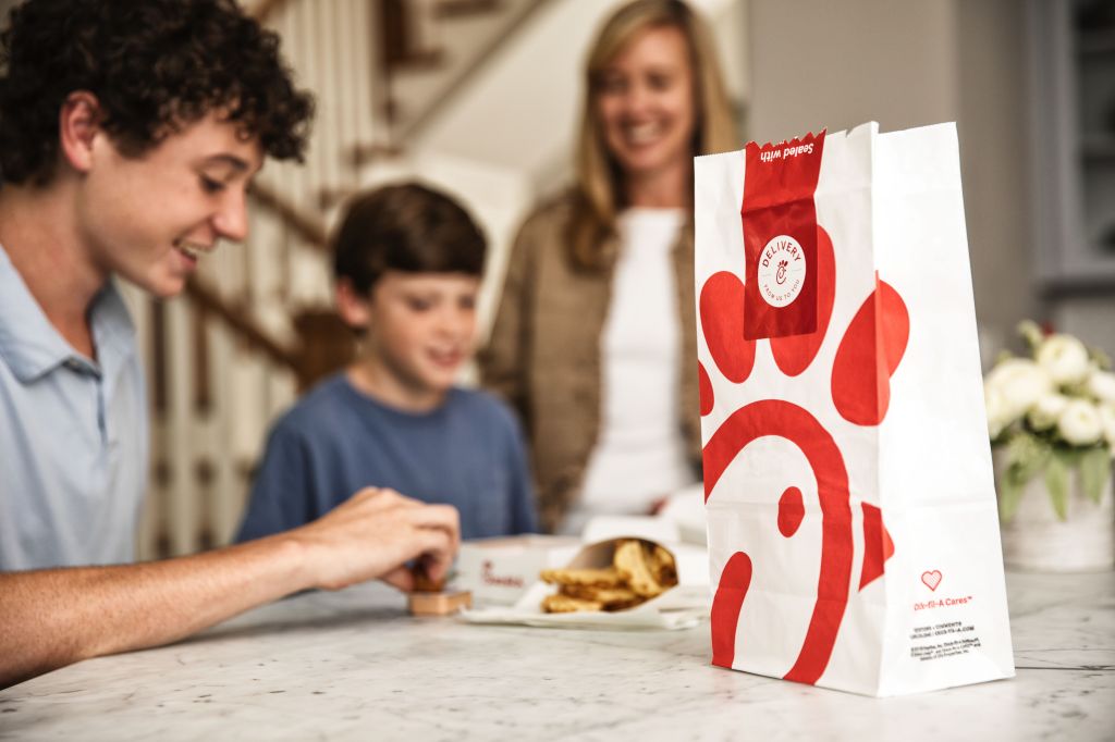 A person sitting at a counter with a bag of Chick-fil-A food, Waffle Fries and dipping a Chick-fil-A Nugget into Chick-fil-A Sauce and two people standing behind him