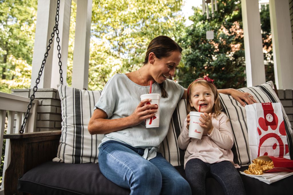 An adult and child sitting on a porch swing, both holding a beverage, with a bag of Chick-fil-A food and Waffle Fries sitting next to them