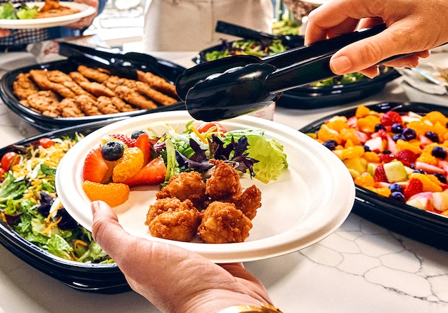 Person holding a plate with Chick-fil-A Nuggets, fruit and a salad in one hand and tongs in the other hand with a Garden Salad Tray, Chocolate Chunk Cookie Tray and a Fruit Tray in the background